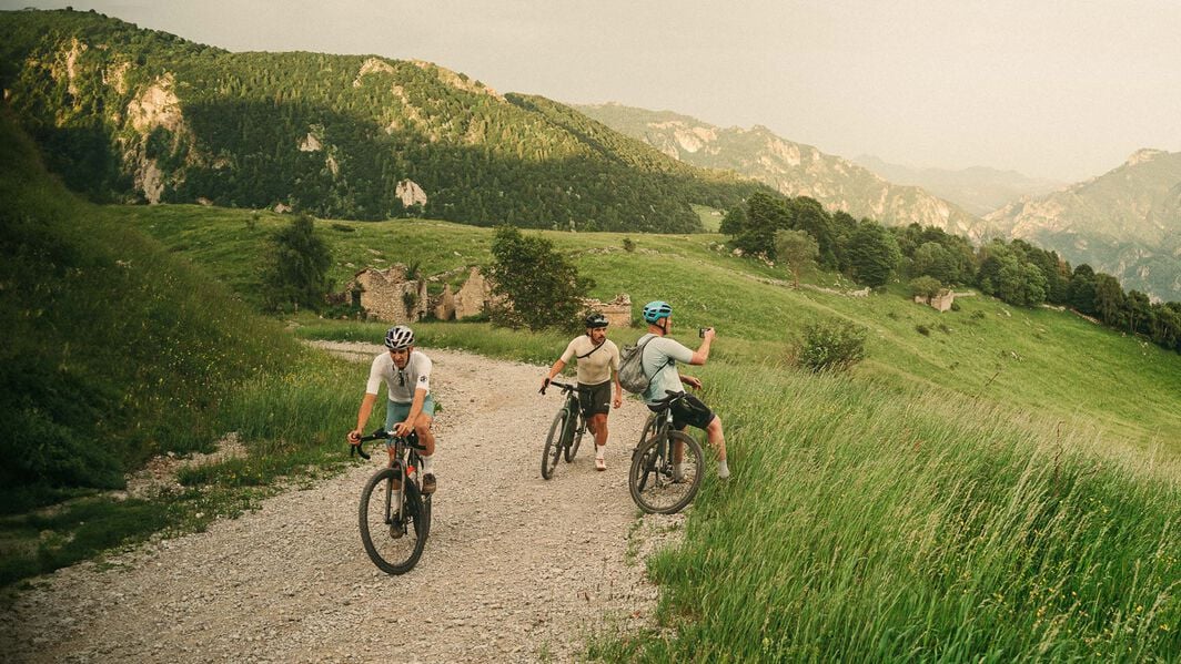 Three cyclists pausing on a gravel mountain trail, surrounded by green hills, while checking directions or planning the next section of their ride.